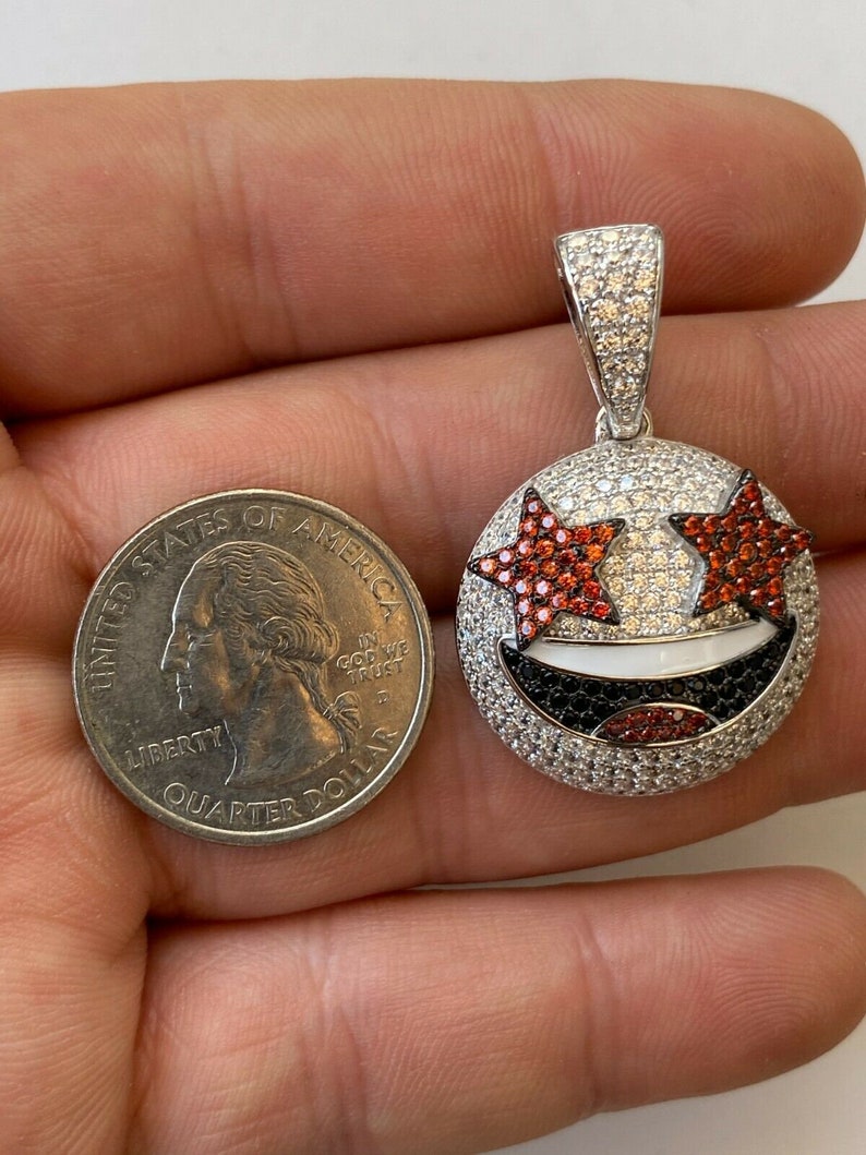 Puede incluir: Un colgante de plata con un dise&ntilde;o de cara sonriente. La cara sonriente tiene ojos en forma de estrella roja, una boca blanca y una nariz negra. El colgante est&aacute; cubierto de peque&ntilde;as piedras preciosas transparentes y rojas.