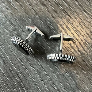 Op de afbeelding: Een paar zilveren manchetknopen met een zwart midden en een getextureerde, rechthoekige rand. De manchetknopen worden gepresenteerd op een donkergrijze, getextureerde ondergrond. De manchetknopen hebben een klassiek, formeel ontwerp.