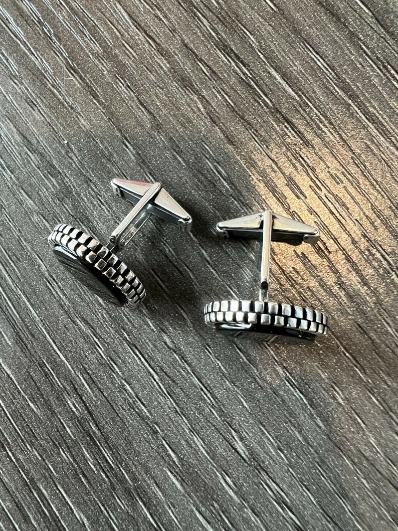 Op de afbeelding: Een paar zilveren manchetknopen met een zwart midden en een getextureerde, rechthoekige rand. De manchetknopen worden gepresenteerd op een donkergrijze, getextureerde ondergrond. De manchetknopen hebben een klassiek, formeel ontwerp.