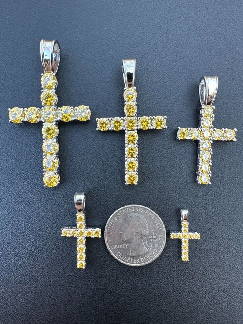 May include: Four silver cross pendants with yellow cubic zirconia stones. The pendants are arranged in order of size, with the largest pendant at the top and the smallest pendant at the bottom. A US quarter is included for size reference.