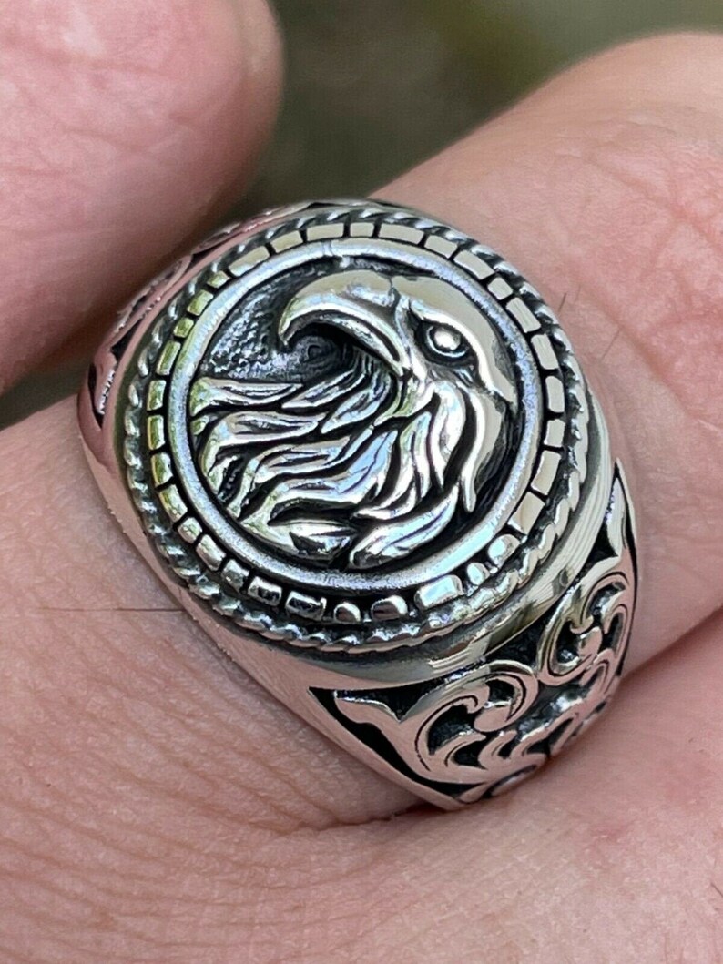 Puede incluir: Un anillo de plata con un dise&ntilde;o de cabeza de &aacute;guila en el centro. El &aacute;guila est&aacute; rodeada por un borde decorativo. La banda del anillo tiene un dise&ntilde;o floral.