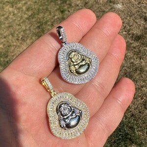 May include: Two pendant necklaces featuring a smiling Buddha design. One pendant has a silver-coloured frame with clear stones and a gold-coloured Buddha. The other pendant has a gold-coloured frame with clear stones and a silver-coloured Buddha.