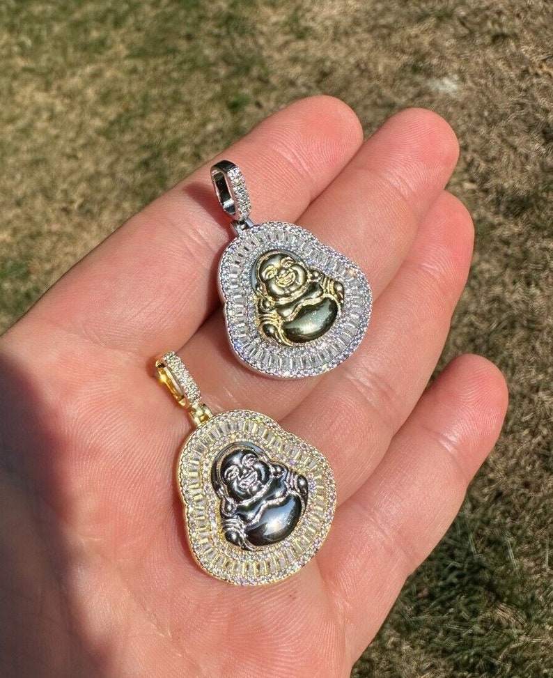May include: Two pendant necklaces featuring a smiling Buddha design. One pendant has a silver-coloured frame with clear stones and a gold-coloured Buddha. The other pendant has a gold-coloured frame with clear stones and a silver-coloured Buddha.