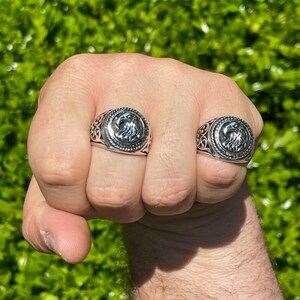 Puede incluir: Dos anillos de plata con un dise&ntilde;o de &aacute;guila en la cara de cada anillo. Los anillos se llevan en el &iacute;ndice y el dedo medio de la mano de una persona.