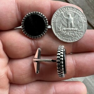 Op de afbeelding: Een paar ronde manchetknopen met een zwart midden en een zilverkleurige, getextureerde rand. Een Amerikaanse kwart dollar munt ligt naast de manchetknopen ter vergelijking van de grootte. De manchetknopen worden vanuit meerdere hoeken getoond.