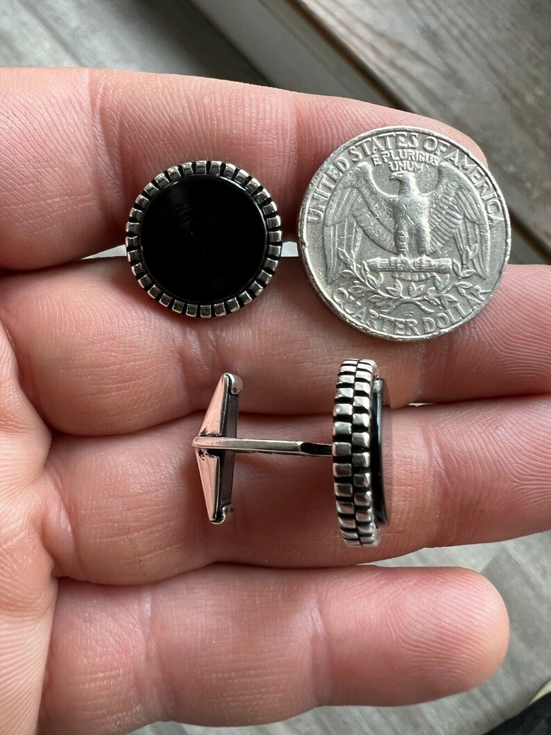 Op de afbeelding: Een paar ronde manchetknopen met een zwart midden en een zilverkleurige, getextureerde rand. Een Amerikaanse kwart dollar munt ligt naast de manchetknopen ter vergelijking van de grootte. De manchetknopen worden vanuit meerdere hoeken getoond.