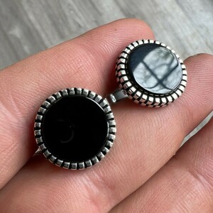 Op de afbeelding: Een paar ronde manchetknopen met een zwart middenstuk en een zilverkleurige, geribbelde rand. De manchetknopen worden gepresenteerd op een lichtgekleurd oppervlak.