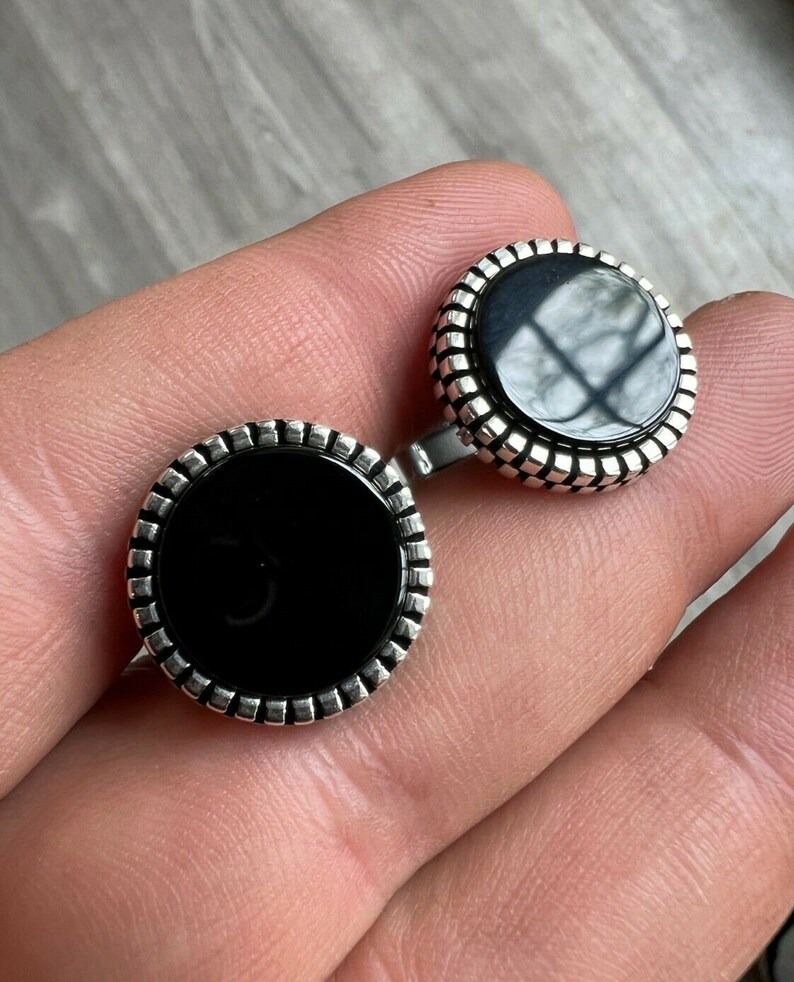 Op de afbeelding: Een paar ronde manchetknopen met een zwart middenstuk en een zilverkleurige, geribbelde rand. De manchetknopen worden gepresenteerd op een lichtgekleurd oppervlak.