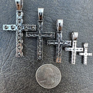 May include: Four silver cross pendants of varying sizes, each adorned with small, clear stones. The crosses are arranged in descending order of size, with the largest cross at the top and the smallest cross at the bottom. A US quarter is included for scale.