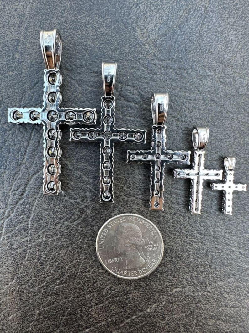 May include: Four silver cross pendants of varying sizes, each adorned with small, clear stones. The crosses are arranged in descending order of size, with the largest cross at the top and the smallest cross at the bottom. A US quarter is included for scale.