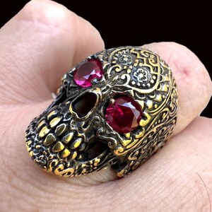 May include: A detailed skull ring with ruby-colored gemstones for eyes. The ring has an antique gold finish and intricate patterns across the skull's surface. The ring is held in a hand against a dark background.