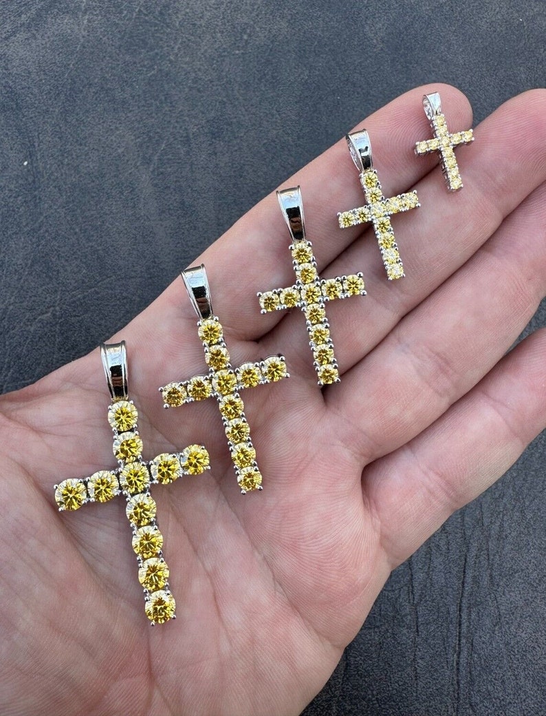 May include: Five silver cross pendants with yellow cubic zirconia stones. The crosses are different sizes, with the largest cross at the bottom and the smallest cross at the top.