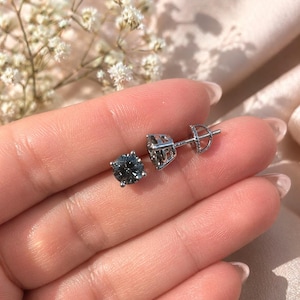 Puede incluir: Un par de pendientes de plata con gemas redondas y grises. Los pendientes están engastados en un engaste de cuatro puntas y los postes tienen un cierre de mariposa. Los pendientes se muestran en la mano de una persona.