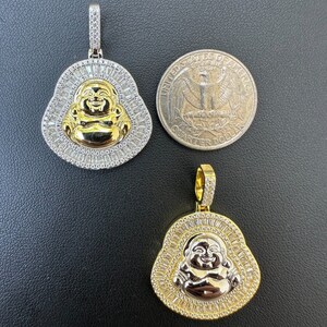 May include: Two gold and silver-tone Buddha pendants. The pendants feature a smiling Buddha face in gold, surrounded by a halo of baguette-cut stones. One pendant is next to a U.S. quarter for size comparison.
