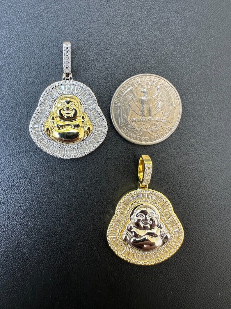 May include: Two gold and silver-tone Buddha pendants. The pendants feature a smiling Buddha face in gold, surrounded by a halo of baguette-cut stones. One pendant is next to a U.S. quarter for size comparison.