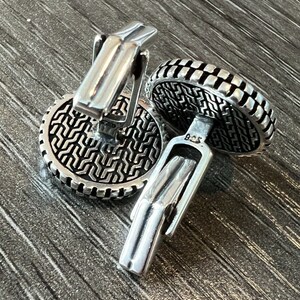 Op de afbeelding: Close-up van twee zilveren manchetknopen. De manchetknopen hebben een cirkelvormig ontwerp met een geometrisch patroon en een tuimelsluiting. Het metaal heeft een gepolijste afwerking en de achtergrond is een gestructureerd grijs oppervlak.