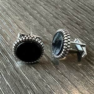 Op de afbeelding: Een paar zilveren manchetknopen met een zwart ovale midden. De manchetknopen hebben een getextureerde, geribbelde rand en een zilveren staaf. De manchetknopen worden gepresenteerd op een houten oppervlak.