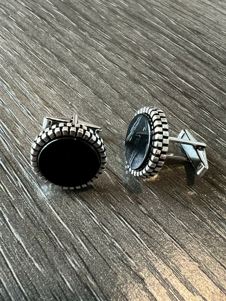 Op de afbeelding: Een paar zilveren manchetknopen met een zwart ovale midden. De manchetknopen hebben een getextureerde, geribbelde rand en een zilveren staaf. De manchetknopen worden gepresenteerd op een houten oppervlak.