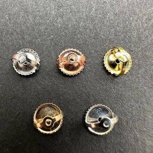 May include: An assortment of five earring backs in various metallic finishes: silver, rose gold, and gold. Each back has a circular design with a textured edge and a loop for securing the earring post. The backs are displayed against a dark background.