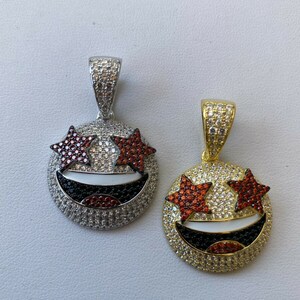 Puede incluir: Dos colgantes de oro y plata con un dise&ntilde;o de cara sonriente. Los ojos son estrellas rojas con piedras blancas y transparentes. La boca es negra con piedras blancas. Los colgantes tienen un aro para sujetar a una cadena.