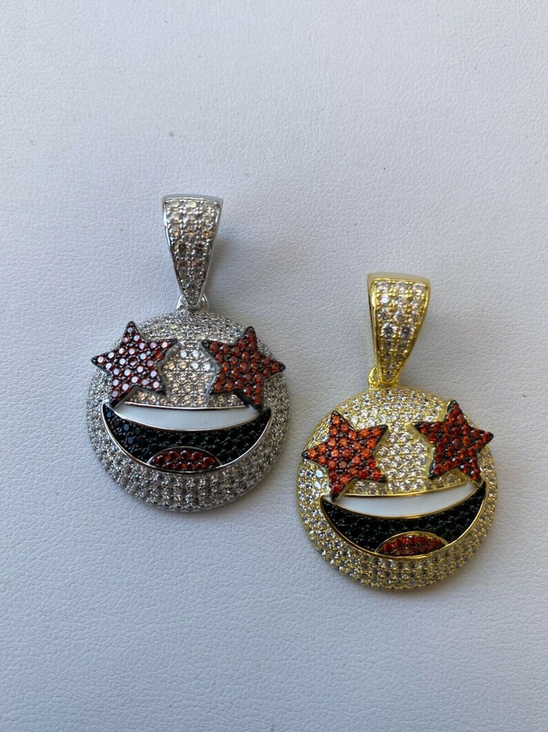 Puede incluir: Dos colgantes de oro y plata con un dise&ntilde;o de cara sonriente. Los ojos son estrellas rojas con piedras blancas y transparentes. La boca es negra con piedras blancas. Los colgantes tienen un aro para sujetar a una cadena.