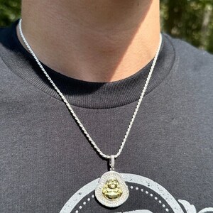 May include: Silver-toned rope chain necklace with a pendant featuring a gold-coloured Buddha figure. The pendant is surrounded by a halo of small, clear stones. The necklace is worn against a dark grey t-shirt.