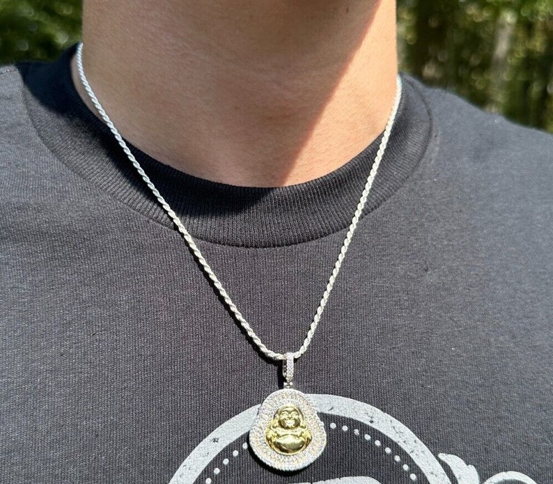 May include: Silver-toned rope chain necklace with a pendant featuring a gold-coloured Buddha figure. The pendant is surrounded by a halo of small, clear stones. The necklace is worn against a dark grey t-shirt.