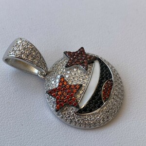 Puede incluir: Un colgante de plata con una luna creciente y dos estrellas engastadas con piedras preciosas rojas y blancas. El colgante est&aacute; rodeado de piedras preciosas blancas m&aacute;s peque&ntilde;as.