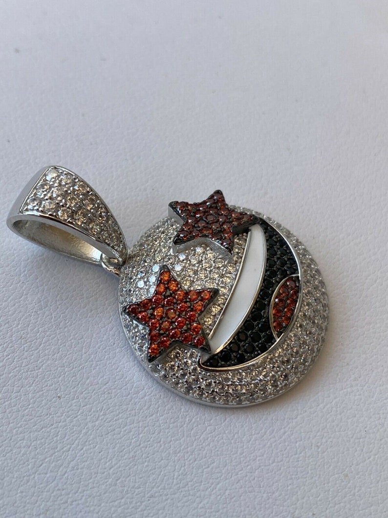 Puede incluir: Un colgante de plata con una luna creciente y dos estrellas engastadas con piedras preciosas rojas y blancas. El colgante est&aacute; rodeado de piedras preciosas blancas m&aacute;s peque&ntilde;as.