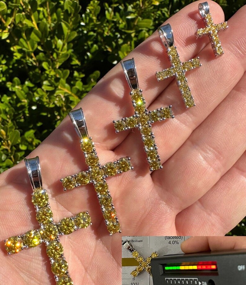 May include: Four silver cross pendants with yellow cubic zirconia stones. The cross pendants are arranged in size order from smallest to largest. The largest cross pendant is about 2 inches tall. The smallest cross pendant is about 1 inch tall. The cross pendants are on a hand.