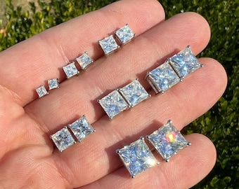 May include: A collection of square-cut, clear gemstone stud earrings in various sizes. The earrings are set in silver-colored metal and are displayed on a hand against a blurred green background. The gemstones sparkle, reflecting light.