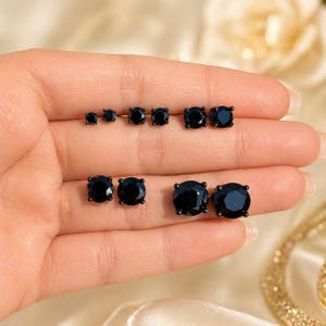 May include: A selection of black stud earrings in different sizes. The earrings have a round shape and a glossy, faceted surface. The earrings are shown on a hand, highlighting the range of sizes.