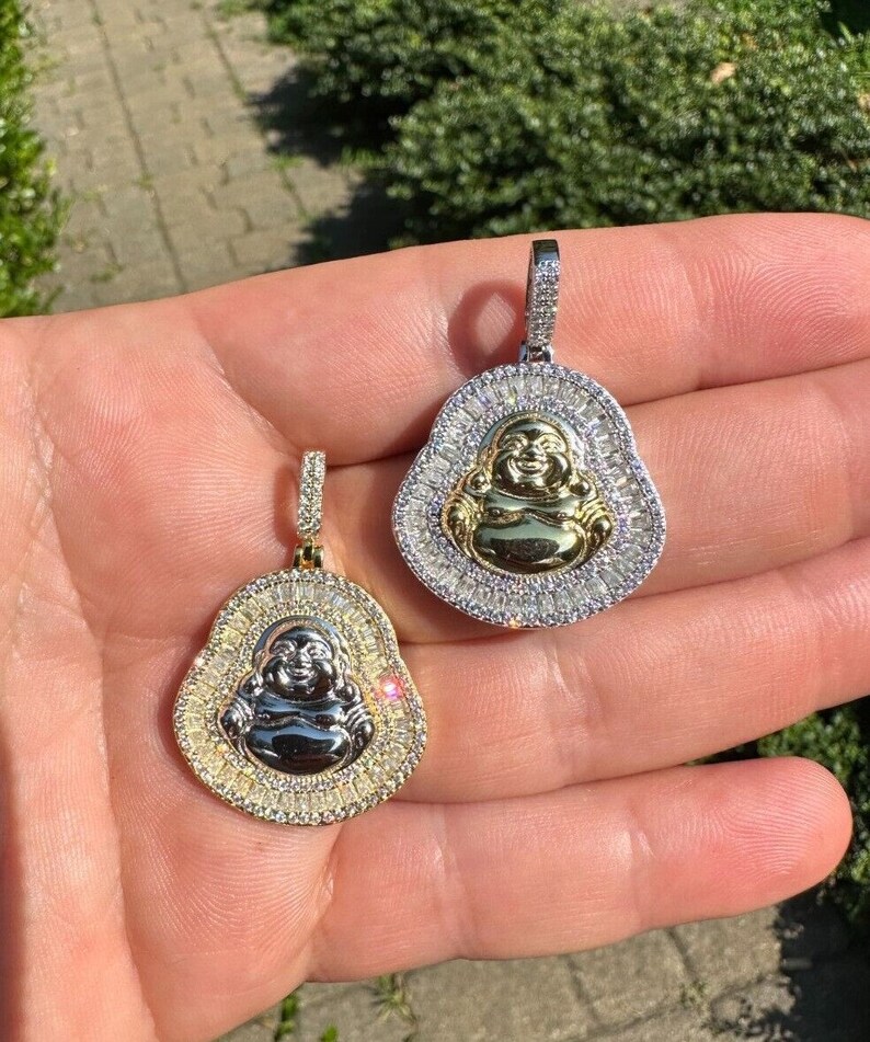 May include: Two gold and silver Buddha pendants. Each pendant features a smiling Buddha figure at the centre, surrounded by a halo of small, sparkling stones. The pendants are held up against a natural background.