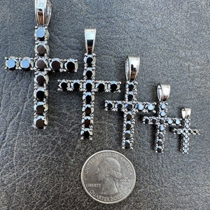 May include: Four silver cross pendants with black cubic zirconia stones. The pendants are arranged in order of size, with the largest pendant at the top and the smallest pendant at the bottom. A US quarter is included for size reference.