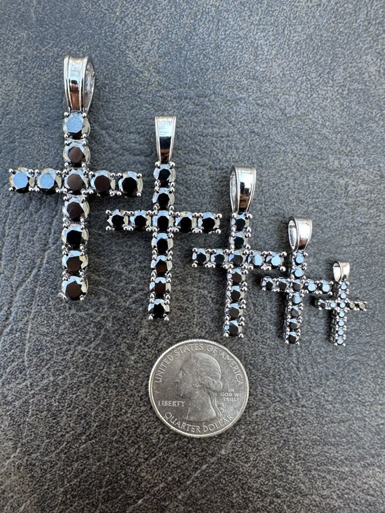 May include: Four silver cross pendants with black cubic zirconia stones. The pendants are arranged in order of size, with the largest pendant at the top and the smallest pendant at the bottom. A US quarter is included for size reference.