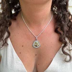 May include: A silver necklace with a pendant depicting a gold Buddha figure. The pendant is framed by a halo of small, clear stones. The necklace is worn against a light-coloured top.
