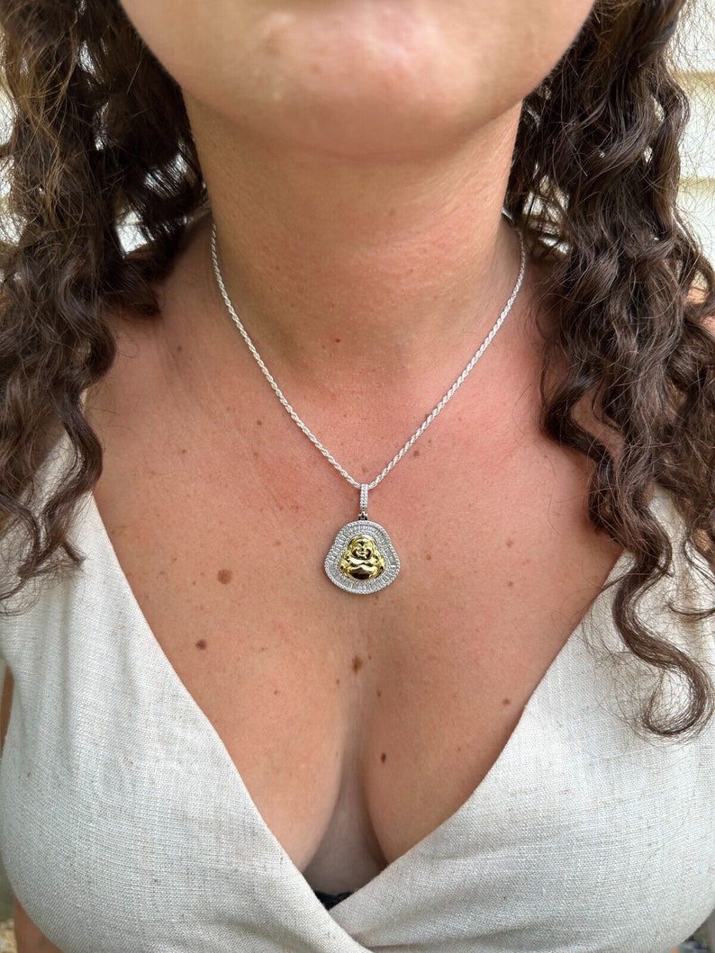 May include: A silver necklace with a pendant depicting a gold Buddha figure. The pendant is framed by a halo of small, clear stones. The necklace is worn against a light-coloured top.