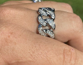Puede incluir: Un anillo plateado con un diseño de eslabones de cadena, incrustado con numerosas piedras preciosas pequeñas y transparentes. El anillo se lleva en un dedo, con un fondo borroso de hierba verde.