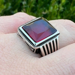 Może przedstawiać: Srebrny pierścionek z dużym, kwadratowym, czerwonym kamieniem szlachetnym. Pierścionek ma szczegółowy, ozdobny wzór.