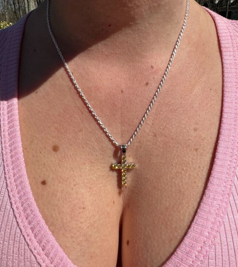 May include: A silver rope chain necklace with a small cross pendant. The cross is set with yellow cubic zirconia stones.