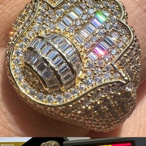 Puede incluir: Un anillo de oro con un gran dise&ntilde;o adornado que presenta una mano de hamsa con un centro redondo. El anillo est&aacute; cubierto de peque&ntilde;os diamantes transparentes.