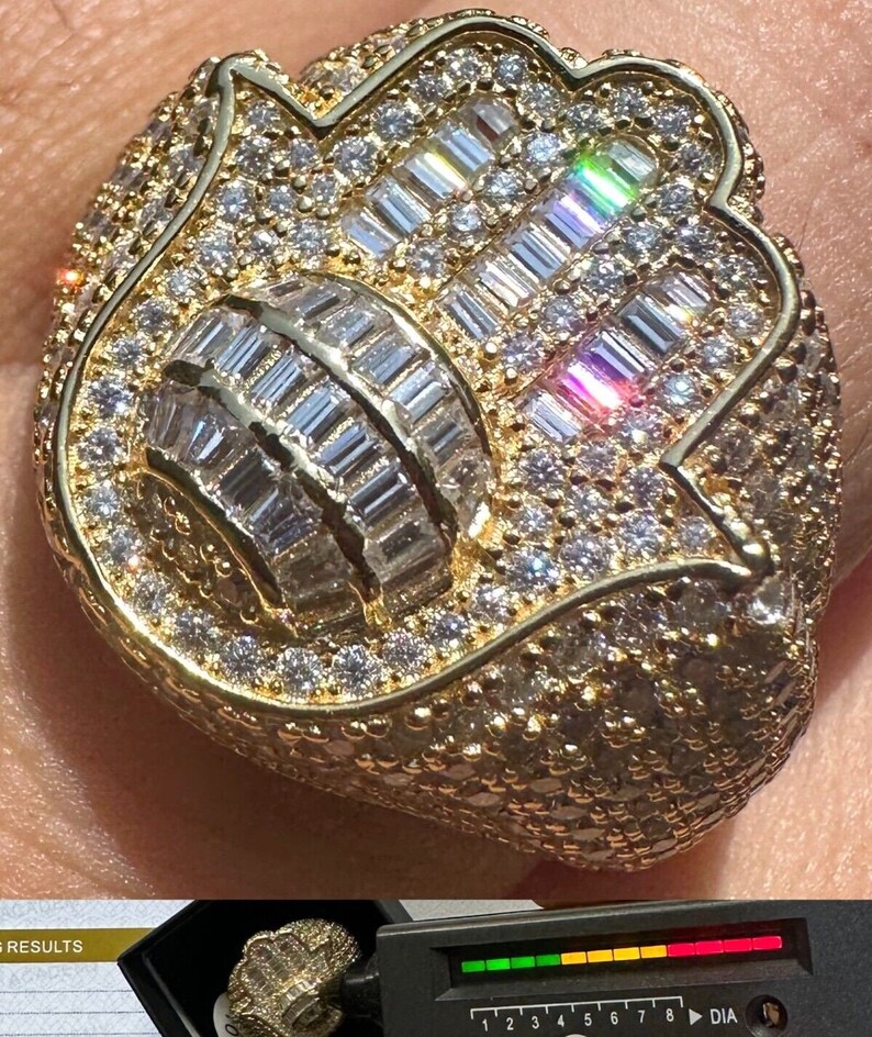 Puede incluir: Un anillo de oro con un gran dise&ntilde;o adornado que presenta una mano de hamsa con un centro redondo. El anillo est&aacute; cubierto de peque&ntilde;os diamantes transparentes.