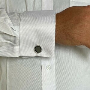 Op de afbeelding: Close-up van een wit overhemd met een manchetknop. De manchetknop heeft een donkergrijze kern met een zilveren kralenrand. Het overhemd is geknoopt en heeft een textuurpatroon.