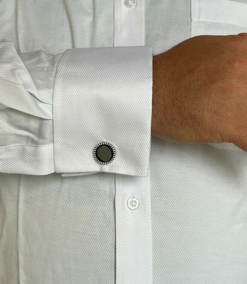 Op de afbeelding: Close-up van een wit overhemd met een manchetknop. De manchetknop heeft een donkergrijze kern met een zilveren kralenrand. Het overhemd is geknoopt en heeft een textuurpatroon.