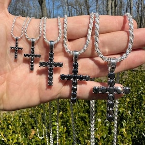 May include: Five silver chains with black diamond cross pendants of varying sizes. The chains are displayed on a hand.