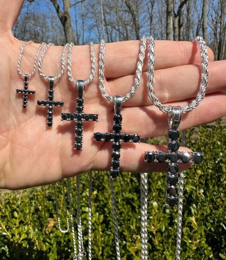 May include: Five silver chains with black diamond cross pendants of varying sizes. The chains are displayed on a hand.