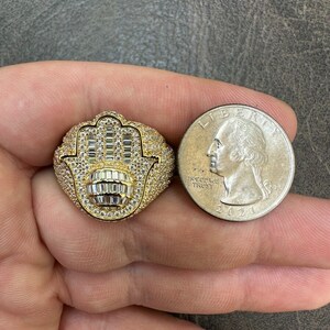 Puede incluir: Un anillo de oro con un dise&ntilde;o de mano de hamsa, engastado con piedras de circonita c&uacute;bica transparentes. El anillo se muestra junto a una moneda de veinticinco centavos de d&oacute;lar estadounidense para comparar el tama&ntilde;o.