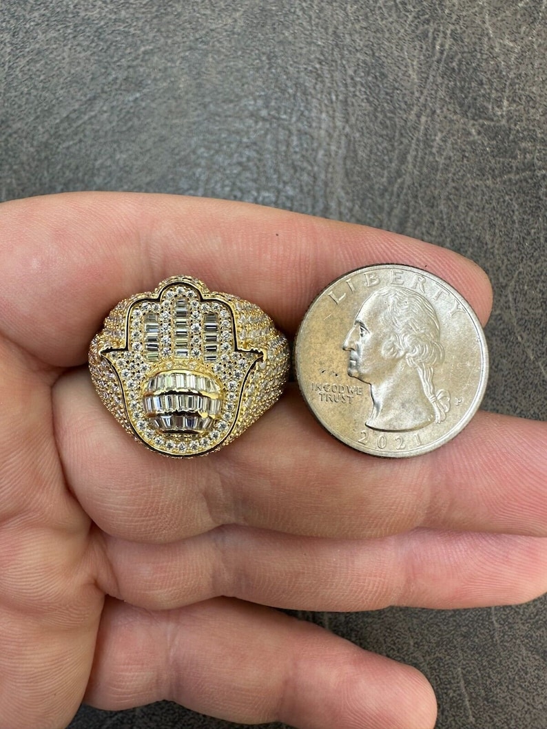 Puede incluir: Un anillo de oro con un dise&ntilde;o de mano de hamsa, engastado con piedras de circonita c&uacute;bica transparentes. El anillo se muestra junto a una moneda de veinticinco centavos de d&oacute;lar estadounidense para comparar el tama&ntilde;o.