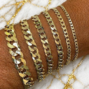 May include: A collection of gold-colored chain bracelets of varying widths displayed on a person's arm. The bracelets have a curb chain design, with links that lay flat. The bracelets are displayed on a surface with gold veining.