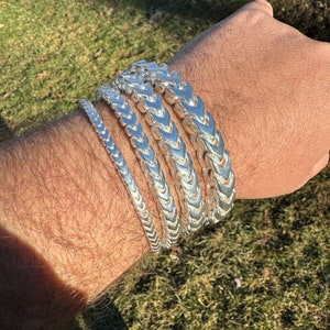 Könnte beinhalten: Ein Set aus vier silbernen Armbändern mit einem Kettenglied-Design. Die Armbänder sind auf dem Handgelenk einer Person gestapelt.